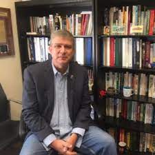 A man in a blazer sits on a chair next to a bookshelf filled with various books. He is looking towards the camera, presenting a relaxed demeanor in an office setting.