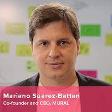 "A man with short brown hair is looking directly at the camera, wearing a black blazer over a pink shirt. The background features blurred colorful sticky notes, suggesting a creative workspace. Text: ""Mariano Suarez-Battan Co-founder and CEO, MURAL"""