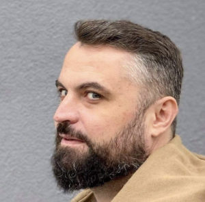 A man with a short beard and styled hair glances sideways with a faint smile on his face. The background is a textured gray wall, providing a neutral context.