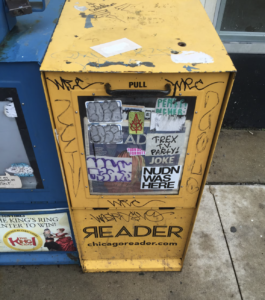 A yellow newspaper vending box stands on a sidewalk, covered in stickers and graffiti. The box is labeled "READER," indicating its contents, amidst a surrounding urban environment.