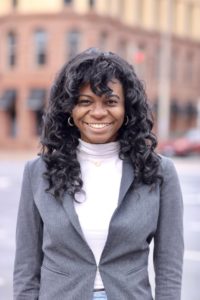 A woman with curly hair smiles brightly while wearing a gray blazer over a white turtleneck. She stands outdoors, with a blurred urban background featuring red traffic lights and buildings.