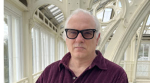 A man with gray hair and black glasses stands confidently indoors, wearing a maroon shirt. Large windows illuminate the space, showcasing a bright, airy environment with intricate architecture.