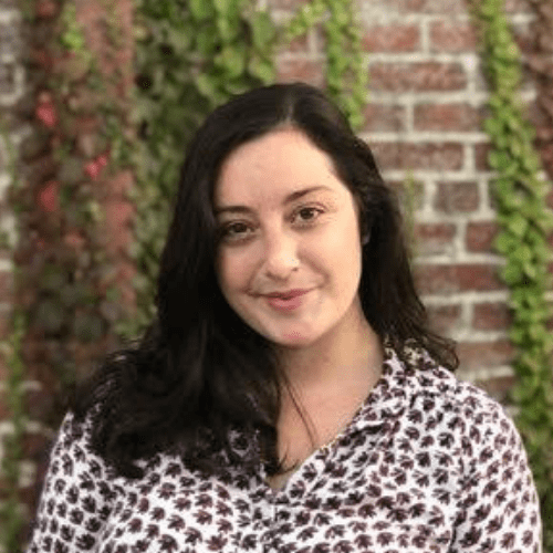 A woman with dark hair smiles while wearing a patterned shirt, standing in front of a brick wall adorned with green vines and foliage.