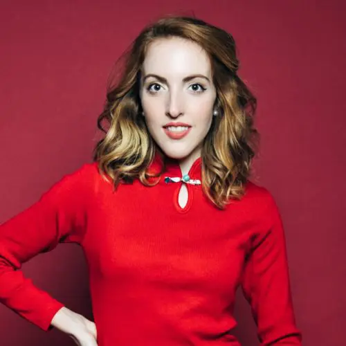 A woman with wavy, shoulder-length hair stands confidently, wearing a bright red sweater. She poses with one hand on her hip against a solid red background.