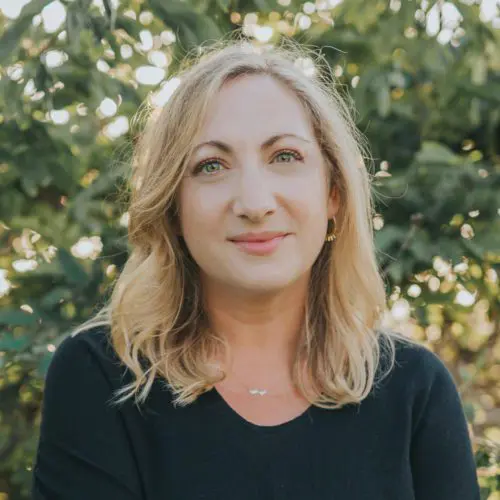 A woman with blonde, wavy hair and green eyes smiles gently. She wears a black top and is surrounded by lush greenery, creating a serene outdoor atmosphere.