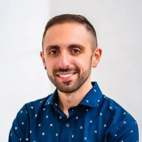 A man with short hair and a beard smiles while wearing a blue shirt with a small patterned design. He is in a well-lit indoor setting with a neutral background.