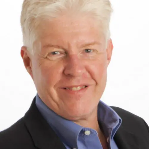 A man with light, slightly tousled hair smiles warmly. He wears a dark blazer over a blue shirt. The background is bright and unobtrusive, emphasizing his friendly expression.