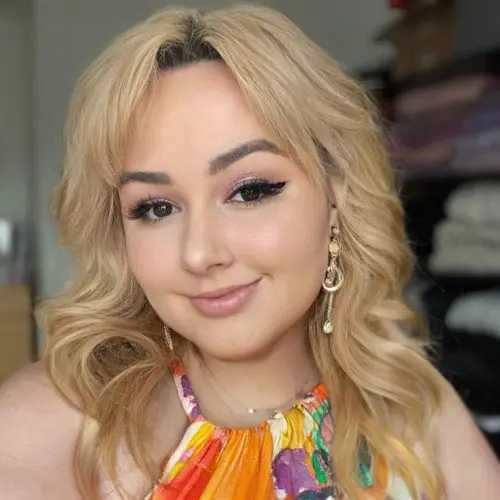 A woman with blonde, wavy hair smiles at the camera, wearing colorful clothing and earrings. She is indoors, with blurred shelves of folded fabric in the background.