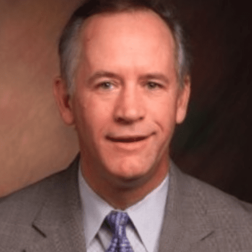 A man dressed in a gray suit and light blue tie smiles for the camera. The background features soft, muted colors that create a professional atmosphere.