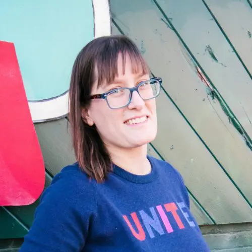 A woman wearing glasses with a blue sweater displaying the word "UNITE" smiles while standing against a background of green wooden panels and a large, colorful sign.