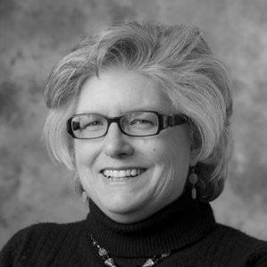 A smiling woman with short, wavy hair wears glasses and a dark turtleneck sweater. She is posed against a blurred, neutral backdrop, conveying a friendly and approachable demeanor.