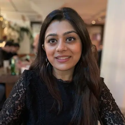 A woman with long, dark hair and hoop earrings sits at a table in a restaurant, smiling at the camera. Soft lighting and people in the background create a cozy atmosphere.