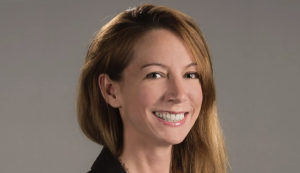 A woman with long brown hair smiles brightly, facing the viewer. She wears a black jacket, set against a soft gray background. Her expression conveys warmth and approachability.
