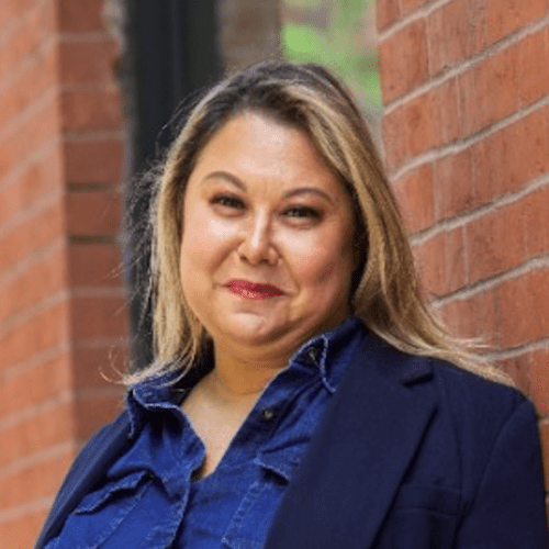 A woman with long, wavy hair smiles warmly, wearing a dark blazer over a blue shirt, leaning against a brick wall in a well-lit outdoor setting.