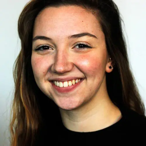 A woman with long, wavy hair smiles brightly, showcasing her teeth. She wears a dark top against a light background, creating a cheerful and engaging expression.