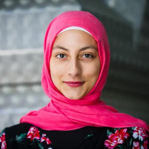 A woman wears a vibrant pink hijab, smiling gently. She stands in front of a textured, neutral background, with floral patterns on her black top.