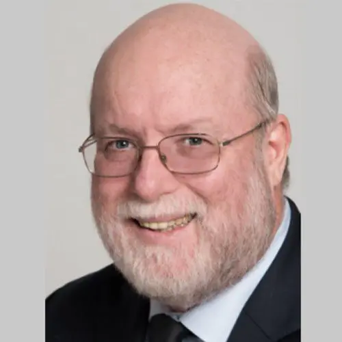 A smiling man with glasses and a beard is looking directly at the viewer. He is dressed in a dark suit, and the background is light gray.