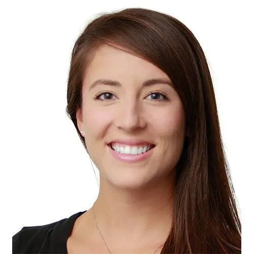 A woman with long brown hair is smiling directly at the viewer. She is wearing a black top, and the background is plain white, enhancing her friendly expression.