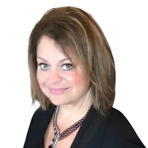 A woman with shoulder-length brown hair smiles lightly, wearing a black top and a necklace with multiple chains, set against a plain white background.