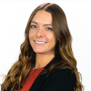 A woman with long, wavy brown hair smiles confidently while posing in a professional setting. She wears a black blazer over a red top, against a plain white background.