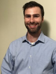 A smiling man with a beard stands in front of a neutral-colored wall, wearing a light blue checkered shirt. His posture is relaxed, conveying a friendly demeanor.