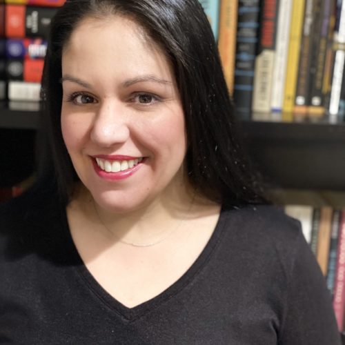 A person with long black hair smiles and wears a black shirt. They stand in front of a bookshelf filled with various books, suggesting a cozy indoor setting.