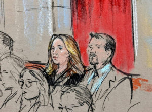 Two individuals sit attentively in a courtroom setting. One has long hair, and the other sports a trimmed beard. The background features red curtains, suggesting a formal atmosphere.