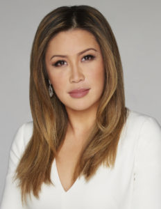 A woman with long, straight, brown hair and wearing a white top is gazing directly at the camera, with a neutral expression, against a light gray background.