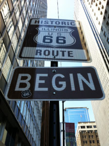 A sign displays "HISTORIC ROUTE 66" above "BEGIN," indicating the start of this famous highway, surrounded by tall buildings in an urban setting.