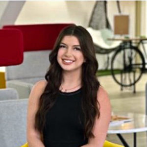A woman with long, dark hair smiles while seated in a colorful, modern lounge. The background features minimalist furniture and a decorative bicycle, creating a vibrant, contemporary atmosphere.