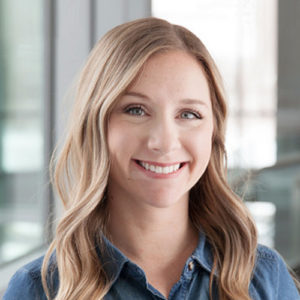 A woman with long, wavy blonde hair smiles warmly. She wears a denim shirt and stands against a backdrop of modern, slightly blurred architectural elements, suggesting a professional environment.