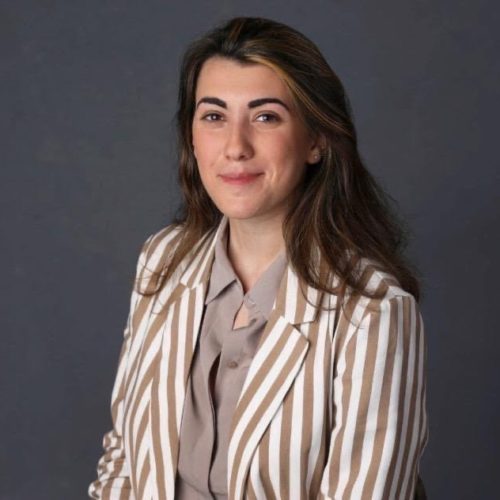 A woman sits against a neutral gray background. She wears a striped blazer over a light shirt, smiling confidently with her hair down, creating a professional and approachable appearance.
