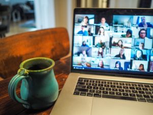 A green ceramic mug filled with dark liquid rests beside a laptop displaying a video conference with numerous participants. The setting features a wooden table and soft interior lighting.