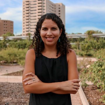 A woman with curly hair stands confidently with her arms crossed in a garden setting, surrounded by greenery and urban buildings in the background under a clear blue sky.
