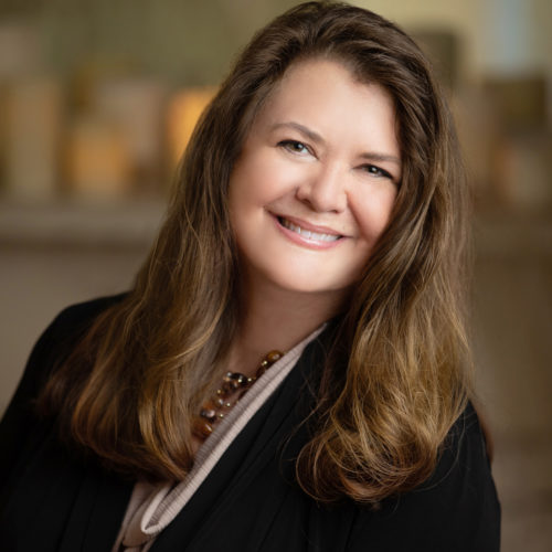 A woman with long, wavy hair smiles warmly. She wears a black jacket and a necklace, posed in a softly lit environment with blurred, illuminated objects in the background.