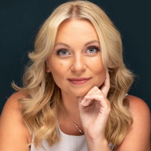 A woman with wavy blonde hair rests her chin on her hand, gazing thoughtfully. The background is dark, enhancing her features and expression. She appears engaged and contemplative.