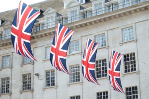 Five Union Jack flags hang from a building, gently swaying in the breeze against a backdrop of light-colored stone walls and large windows, suggesting a festive or patriotic atmosphere.