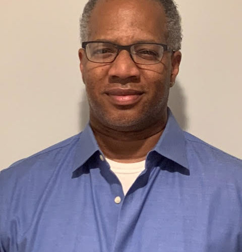 A man wearing a blue collared shirt over a white t-shirt stands against a plain light-colored background, looking directly at the viewer with a neutral expression.