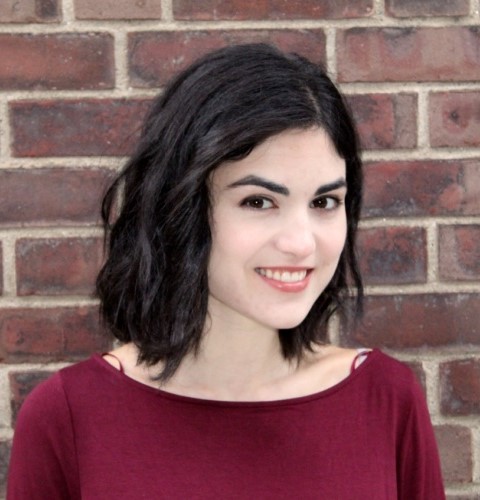 A woman with dark, wavy hair smiles while facing the camera. She is wearing a maroon top, standing against a textured brick wall.