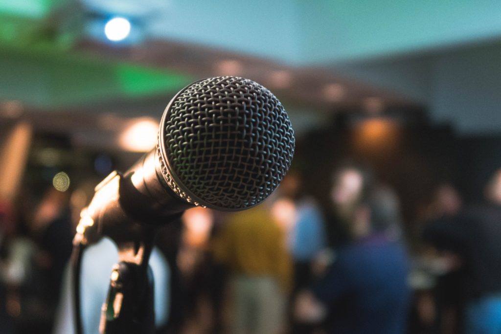 A microphone stands prominently in the foreground, ready for use, while a blurred crowd engages in conversation in a softly lit, modern environment.