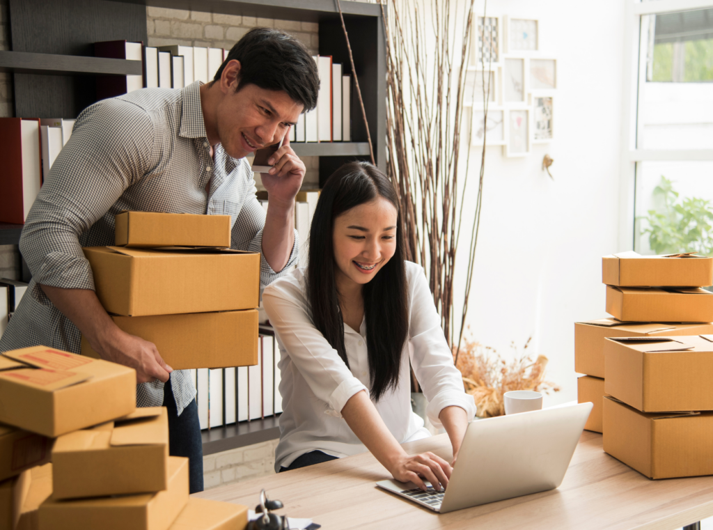Two small business owners packing orders, showcasing teamwork and dedication in their thriving enterprise. The man stands on the phone overlooking a woman's shoulder as she types. This duo seeks to strengthen PR efforts in their small business. 